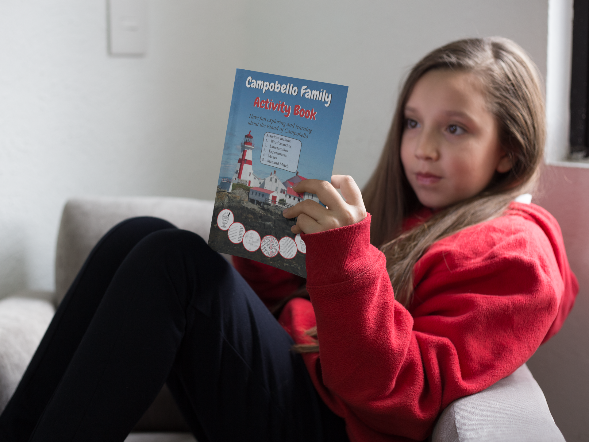 A girl sits on a couch reading the Campobello Family Activity Book